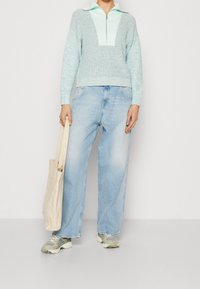 Hellblauer Strickpullover mit V-Ausschnitt und Reißverschluss, kombiniert mit hellblauen, weiten Jeans und grau-grünen Turnschuhen, hält eine beige Tragetasche.