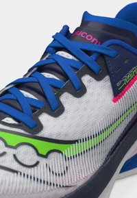 Κοντινή λήψη ενός λευκού και ναυτικού παπουτσιού τρεξίματος με μπλε κορδόνια, νέον πράσινες και ροζ λεπτομέρειες, και επάνω μέρος από δίχτυ.