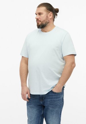 Mężczyzna z brodą i związanymi do tyłu włosami stoi z jedną ręką w kieszeni, ubrany w jasnoniebieski t-shirt i niebieskie dżinsy na białym tle.
