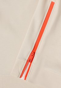 Gros plan sur un tissu beige avec une fermeture éclair orange vif verticale partiellement dézippée montrant le curseur et les détails de la couture.