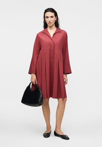 Γυναίκα που φοράει μακρυμάνικο, κόκκινο φόρεμα με κουμπιά, κρατάει μια μαύρη τσάντα χειρός και φοράει μαύρα, επίπεδα παπούτσια, στέκεται μπροστά σε απλό φόντο.