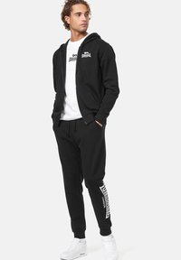 Zwarte zip-up hoodie gecombineerd met zwarte joggers met een witte logoprint aan de zijkant. gedragen met een wit T-shirt en witte sneakers.