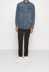 Mann in gebuttonierter blauer Jeansjacke, schwarzen Hosen und weißen Sneakers steht vor einem schlichten Hintergrund.