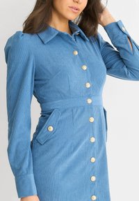 Blue corduroy dress with long sleeves, a collar, side pockets, and gold buttons down the front. Textured fabric with vertical stripes.