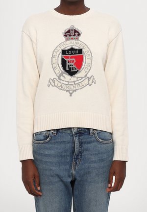Pull en tricot crème avec un logo écusson rouge et noir accompagné de texte, porté avec un jean bleu délavé moyen sur une personne debout.