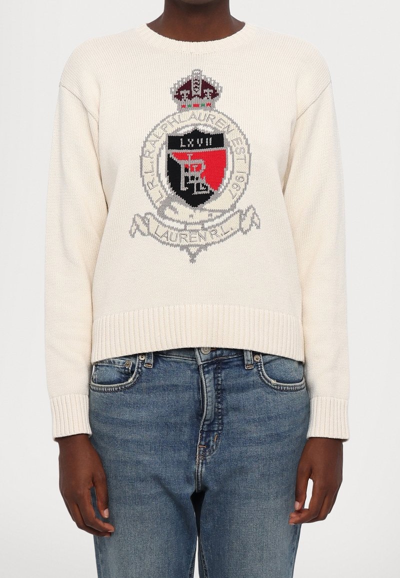 Pull en tricot crème avec un logo écusson rouge et noir accompagné de texte, porté avec un jean bleu délavé moyen sur une personne debout.