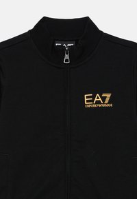 Čierna zipsová bunda vyrobená z mäkkého materiálu, s vysokým golierom, bočnými vreckami a zlatým logom na hrudi. Hladká textúra po celom povrchu.