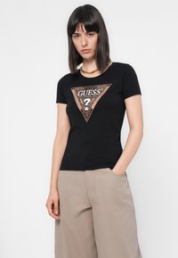 Kvinde med glat, mørkt hår iført en sort Guess-logo T-shirt og beige løse bukser, stående med den ene hånd i lommen mod en hvid baggrund.