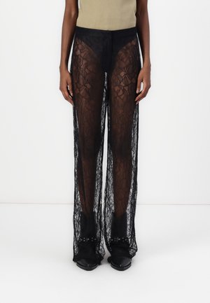 Persona con pantalones acampanados de encaje negro translúcido sobre pantalones cortos negros y botas negras con tachuelas, de pie contra un fondo blanco.