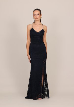 Mujer con un vestido largo negro de encaje con tirantes finos y una abertura al muslo, de pie frente a un fondo beige liso.
