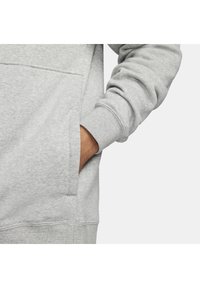 Ruka v přední kapse světle šedého svetr s ribbed manžetami, zobrazeno na neutrálním pozadí.