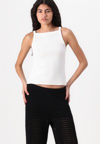 Top blanco sin mangas con escote cuadrado, textura acanalada, combinado con pantalones de punto negros que presentan un diseño de tejido abierto y cinturilla elástica.