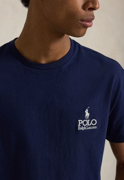 Νέο άτομο που φοράει μπλε μπλουζάκι Polo Ralph Lauren με λευκό κεντημένο λογότυπο στο αριστερό στήθος, στραμμένο ελαφρώς προς τα δεξιά.