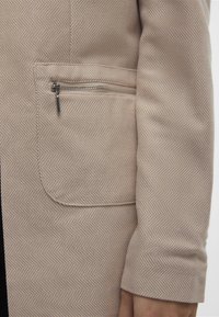 Chaqueta de color beige con textura que presenta un patrón de espiga y un bolsillo con cremallera en el lado frontal izquierdo. Tejido suave con mangas ajustadas.