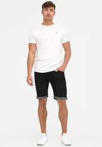 Weißes T-Shirt mit Rundhalsausschnitt und kleinem Logo, kombiniert mit schwarzen Jeansshorts, die am Saum umgekrempelt sind. Weiße Sneaker vervollständigen den Look.