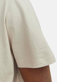Beige Baumwoll-T-Shirt mit kurzen Ärmeln. Glatte Textur und gesäumter Rand, der den Arm in einer Nahaufnahme zeigt.