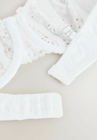 Białe koronkowe ramiączka do biustonosza z regulowanym zapięciem i zapięciami na haftki na jasnym neutralnym tle.