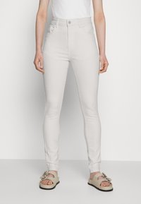 Jeans skinny brancos de cintura alta com quatro bolsos e fecho de botão, combinados com sandálias deslizantes bege com decoração em metal.