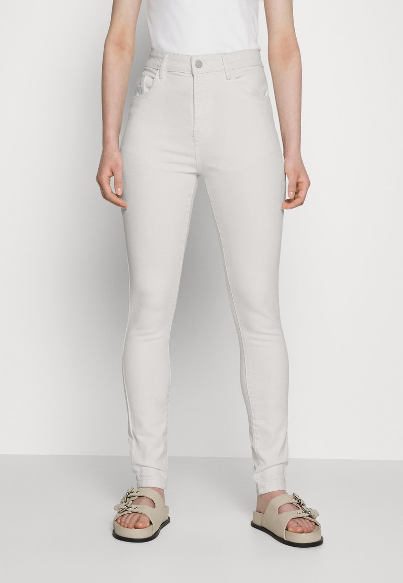 Jeans skinny brancos de cintura alta com quatro bolsos e fecho de botão, combinados com sandálias deslizantes bege com decoração em metal.