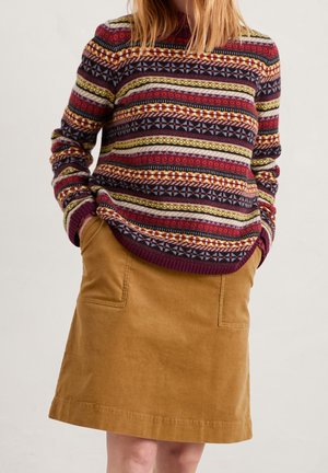 Femme portant un pull multicolore à motifs et une jupe en velours côtelé marron avec des poches, les mains glissées dans les poches de la jupe.