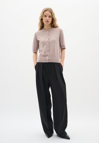 Hellrosa Kurzarm-Cardigan mit Rundhalsausschnitt und vorderen Knöpfen, kombiniert mit schwarz hohen Hosen und schwarzen, spitzen Schuhen.