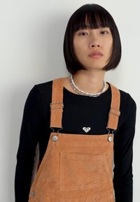 Person with short black hair wearing a black long-sleeve shirt, beige corduroy overalls, and a silver beaded necklace, against a plain background.