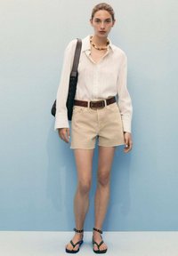 Chemise blanche à rayures verticales légères, shorts beiges, ceinture marron, collier de perles et sac noir à bandoulière, associés à des sandales plates.