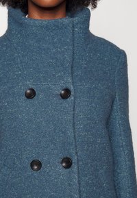 Modrý vlněný kabát se vysokým límcem, dvojitým zapínáním a dvěma černými knoflíky. Textura je měkká s jemným melírovaným vzorem.