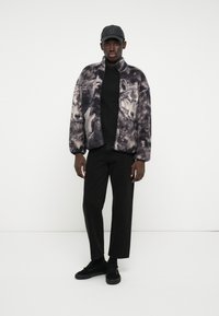 Ψεύτικο γούνινο μπουφάν με εκτυπώσεις από γκρι και μαύρους λύκους, συνδυασμένο με μαύρη ζιβάγκο, μαύρο παντελόνι και μαύρα αθλητικά παπούτσια. Μαύρο καπέλο ολοκληρώνει το σύνολο.