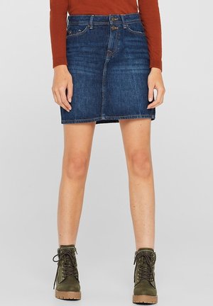 Vrouw met een blauwe denim minirok, een roestrode top met lange mouwen en groene veterlaarzen tot aan de enkels, staand op een neutrale achtergrond.