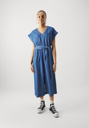 Denim jumpsuit met korte mouwen, V-hals, ceintuurlijn en cropped wijde benen. Draagt met zwarte hoge sneakers en witte sokken.