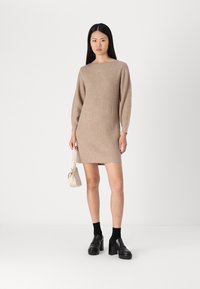 Robe tricotée beige avec une texture côtelée, un encolure bateau et des manches longues. Portée avec des chaussures plates-formes noires épaisses et un petit sac blanc.