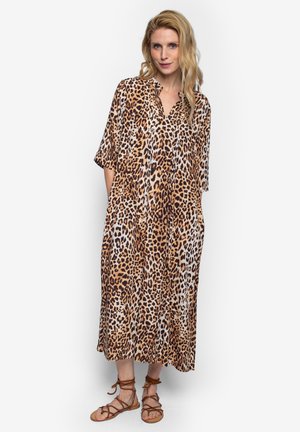 Blonde Frau trägt ein locker sitzendes, midi langes Kleid mit Leopardenmuster und ellbogenlangen Ärmeln sowie braune flache Sandalen mit Schnürung, steht vor einem weißen Hintergrund.