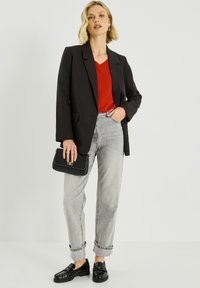 Blazer negro sobre una camiseta roja, combinado con jeans grises remangados y mocasines negros de charol. Sosteniendo un bolso negro con textura.