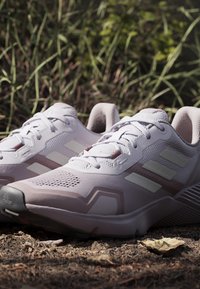 Paar weiße und hellrosa Adidas Laufschuhe auf trockenem Boden mit grünem Gras im Hintergrund.
