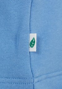 Svijetloplavi pleteni materijal s bijelom etiketom koja sadrži logo zelenog lista. Tekstura je glatka s vidljivim detaljima šivanja.