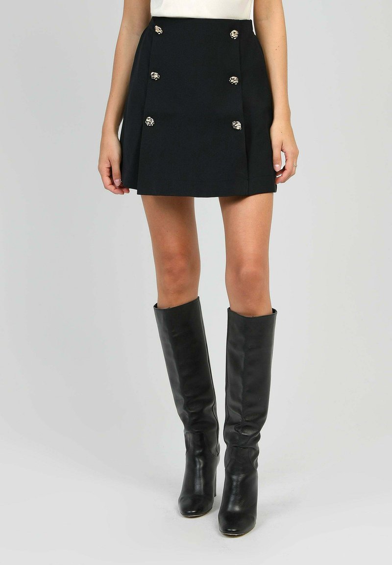 Femme portant une mini-jupe noire avec des boutons décoratifs et des bottes en cuir noir jusqu'aux genoux, se tenant devant un fond uni.