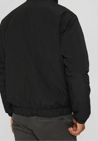 Giubbotto bomber nero con una texture liscia, polsini e orlo elastici, e un colletto alto, con un'etichetta del marchio discreta sul retro.