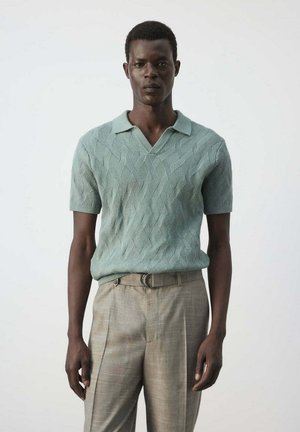 Hombre con camisa polo de punto de manga corta de color verde claro y pantalones beige de talle alto, de pie frente a un fondo liso.