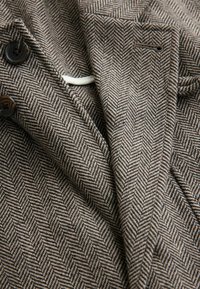 Brown herringbone patterned fabric with two dark buttons on a collar, showing texture and layered folds of the material.
