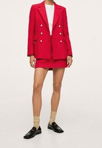 Blazer rojo texturizado con diseño de doble botonadura y botones plateados, combinado con una falda corta a juego. Mocasines negros y calcetines beiges.