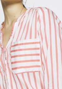 Camicia leggera a maniche corte in cotone con strisce verticali rosa e bianche. Presenta una chiusura con bottoni sul davanti e una tasca sul lato sinistro.