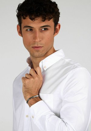 Camicia bianca con colletto e taschino sul petto. Modello che indossa un bracciale d'argento, con maniche corte e una trama di tessuto liscia.