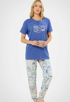 Femme portant un t-shirt bleu à manches courtes avec l'inscription « rêve de la mer » et un pantalon à motifs, tenant une tasse beige sur fond blanc.
