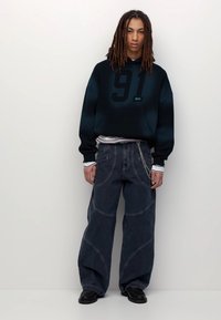 Giovane con dreadlocks che indossa un hoodie nero con il numero 91, jeans larghi scuri con catena e scarpe nere, in piedi contro uno sfondo semplice.