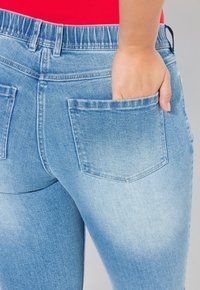 Vaqueros de mezclilla azul claro con una textura desgastada, que cuentan con dos bolsillos traseros y una cinturilla de altura media. Una mano está colocada en un bolsillo.