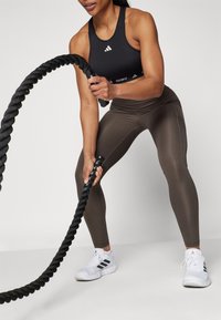 Black athletic crop top, brown leggings, and white shoes. Person grips a black battle rope, showcasing active workout posture.