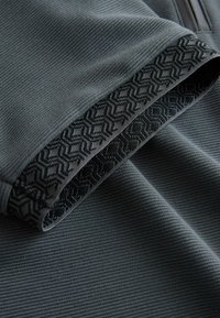 Nærbillede af mørkegråt stof med subtil ribbet struktur og en foldet kant med sort geometrisk mønstret kantbånd.