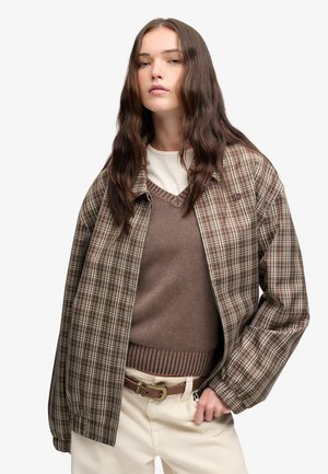 Joven mujer con cabello largo castaño que lleva una chaqueta marrón a cuadros, chaleco de punto marrón, camisa blanca, pantalones beige y un cinturón marrón.