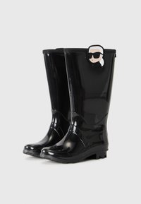 Bottes de pluie noires brillantes avec un visage de dessin animé portant des lunettes de soleil et une queue de cheval blanche attachée près du haut sur un fond blanc uni.